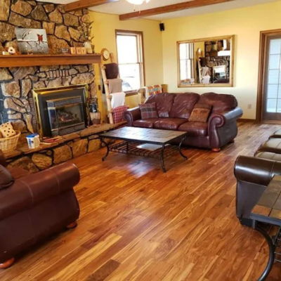 Cozy living room with a stone fireplace, warm wood floors, leather sofas and a coffee table, a ceiling light, and large windows with curtains, viewed from the doorway.